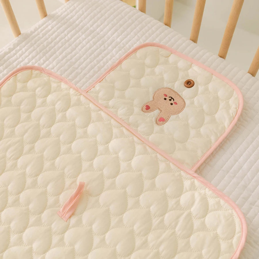 A beige quilted portable diaper changing pad with a cute brown bear design resting on a white textured surface.