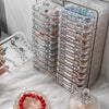 A clear jewelry organizer box filled with multiple stacked compartments and loose beads, resting on a white fluffy surface.