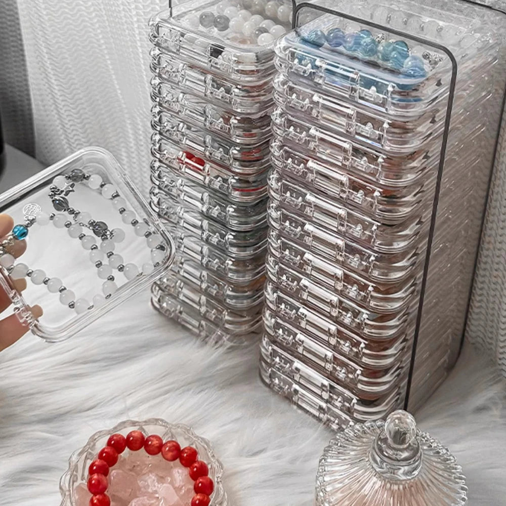 A clear jewelry organizer box filled with multiple stacked compartments and loose beads, resting on a white fluffy surface.