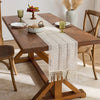 A beige hand-woven boho table runner with tassels is placed on a wooden dining table.