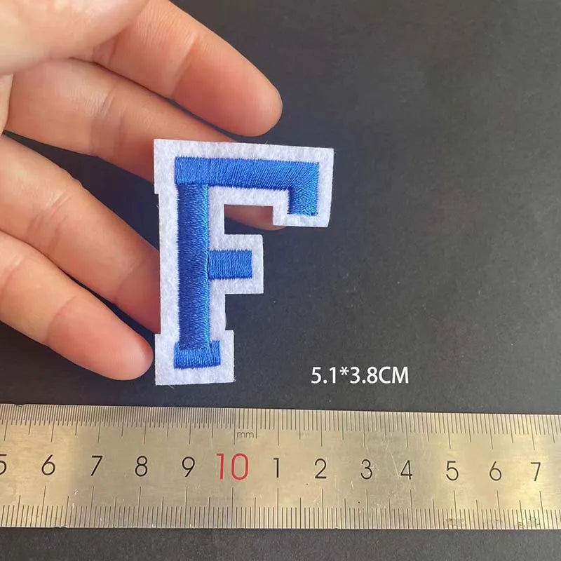 A hand holds a blue embroidered letter F patch measuring 5.1 by 3.8 cm next to a ruler.