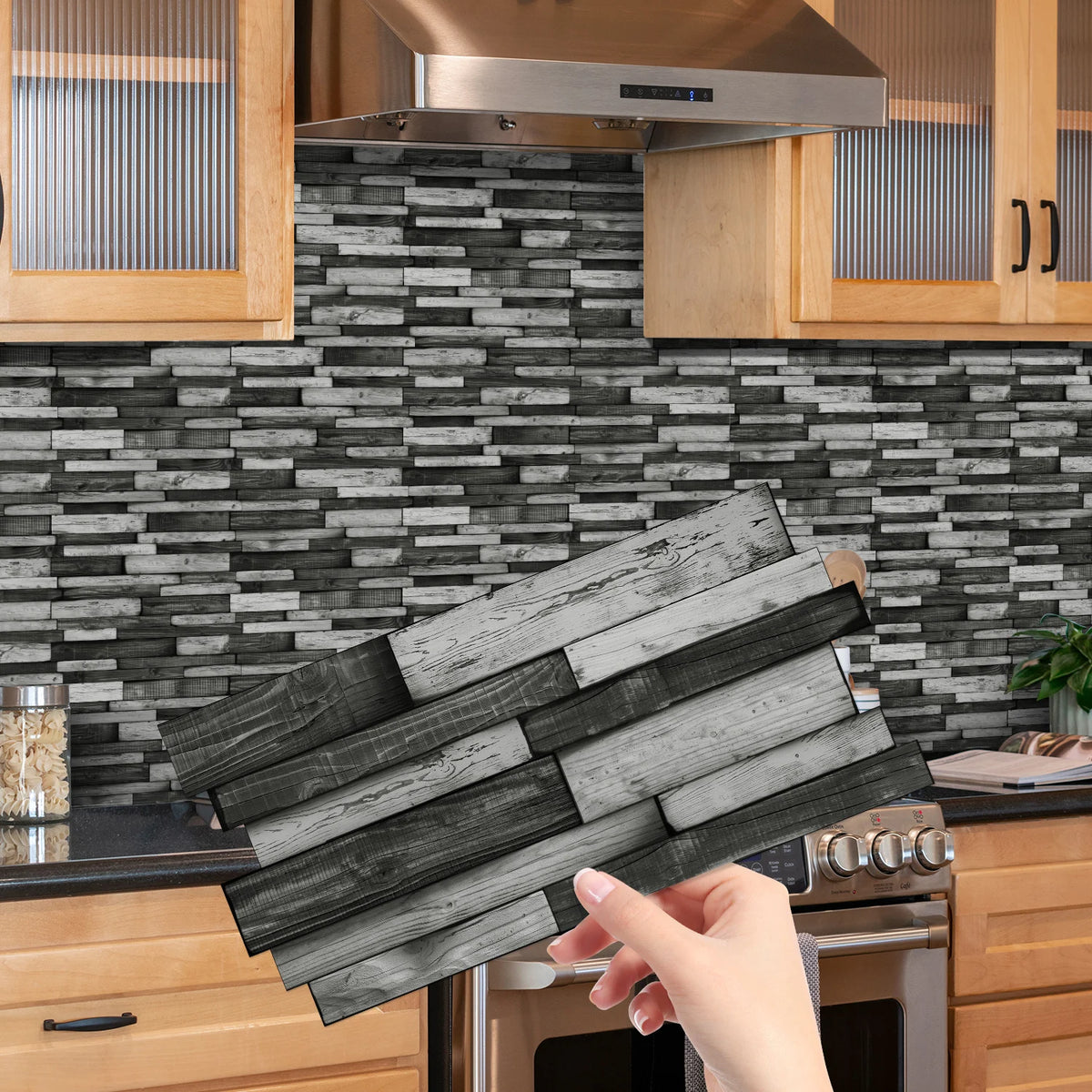 A hand holds a rectangular piece of dark gray wood tile stickers against a textured gray subway tile backsplash in a kitchen 