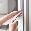 Eine Hand appliziert wiederverwendbares graues PU-Schaumdichtungsband auf einen Fensterrahmen.
