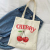 A canvas tote bag with a red cherry print and the text Happy Have a Nice Day, placed on a white sheet next to denim jeans.