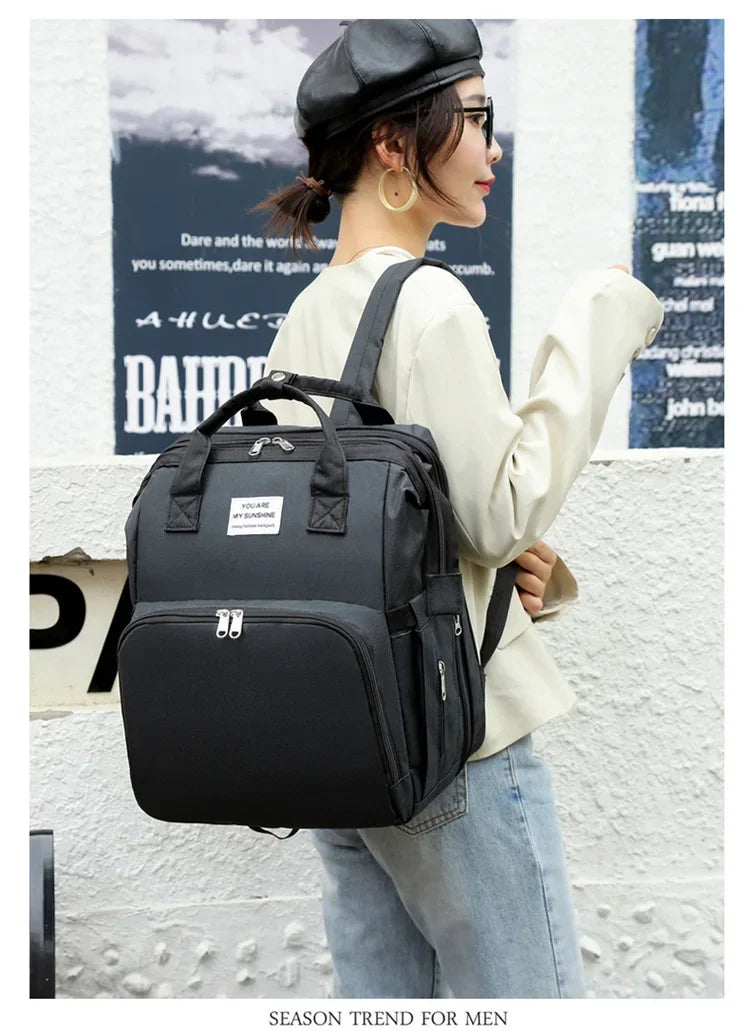 A woman wearing a cream top and jeans carries a black large capacity diaper bag backpack.