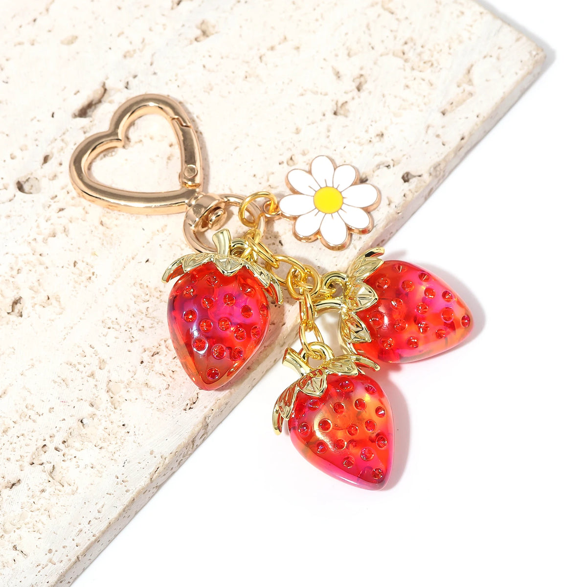 Clochette de clé en forme de fraise ton or avec un détail de fleur blanche sur fond clair.