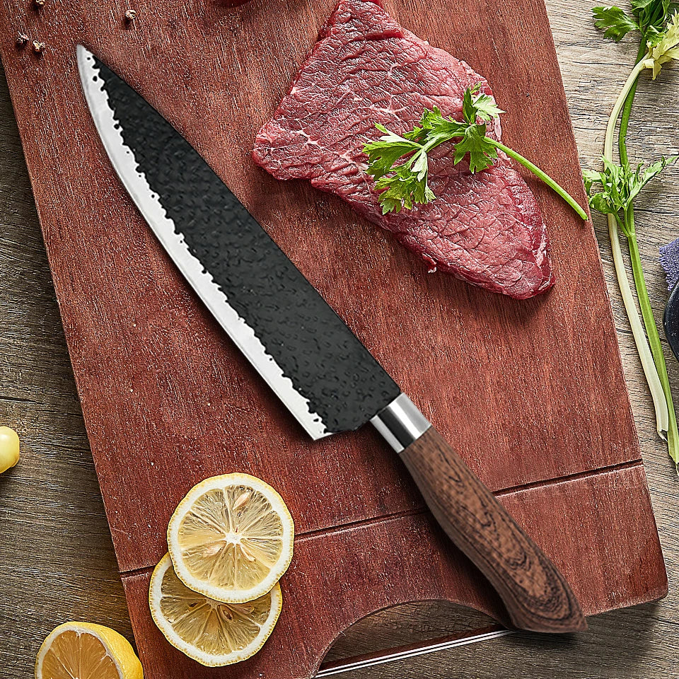A forged hammer-pattern kitchen knife set with a dark blade and wooden handle rests on a cutting board next to a steak and le