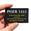 Black embroidered patch featuring Psalm 144:1 text on a fabric tag being held by a hand.