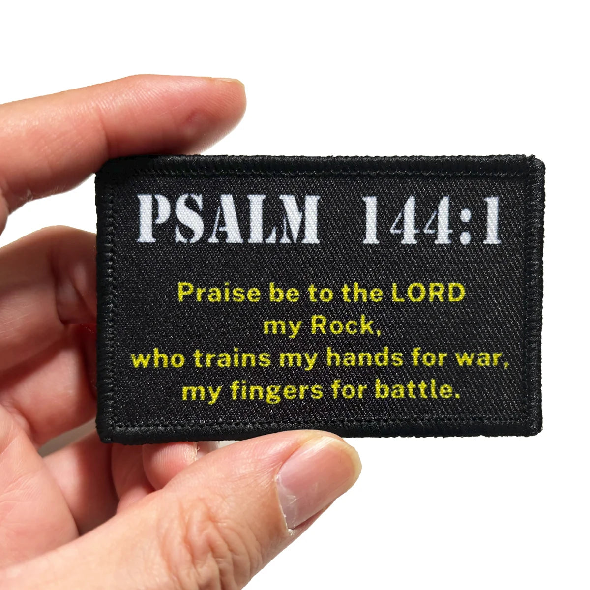 Black embroidered patch featuring Psalm 144:1 text on a fabric tag being held by a hand.