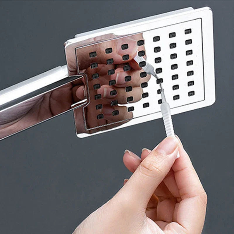 A hand is using a shower cleaning brush set with an anti-clogging nozzle cleaner on a white plastic grid.