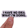 A hand holds a black embroidered iron-on patch with white text that reads I HAVE NO IDEA WHAT I'M DOING.