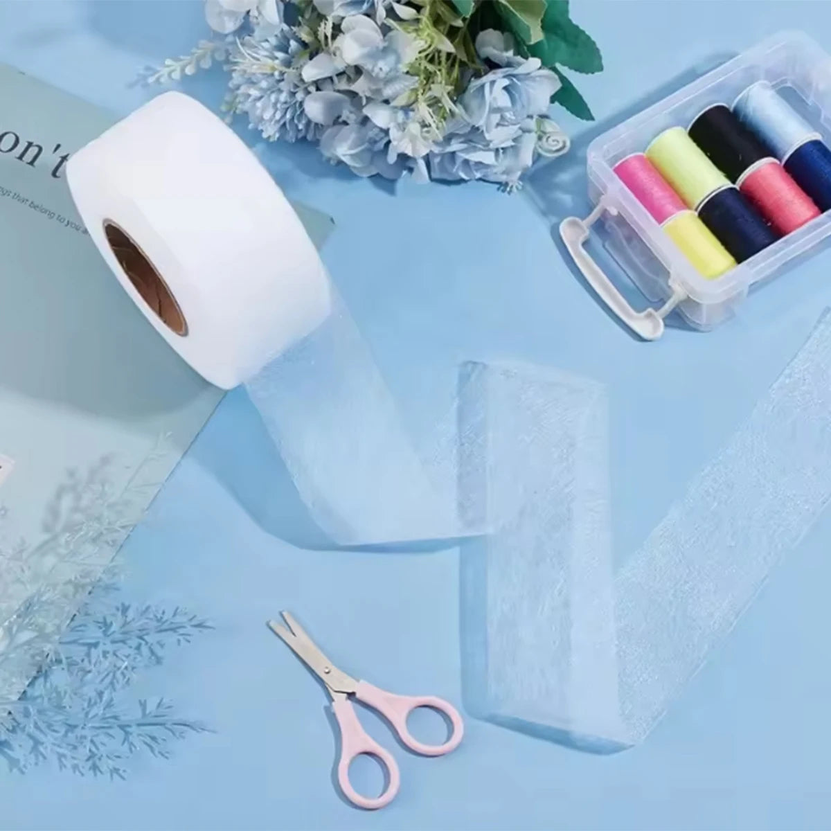 Roll of white no-sew iron-on hem tape next to floral decorations and a small plastic container of colored tapes.