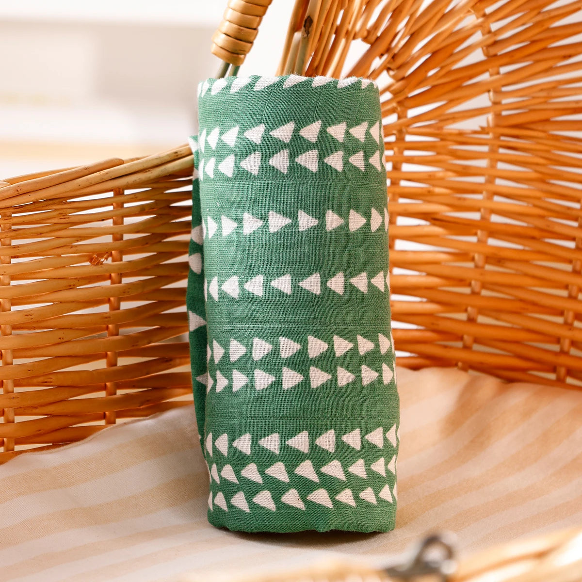 A green muslin baby swaddle blanket with white triangles is displayed next to a woven basket.