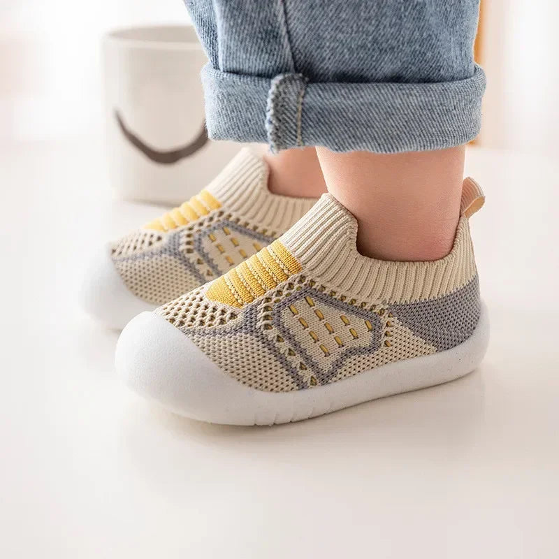 A pair of beige and yellow breathable soft-sole baby shoes with a textured knit upper are shown on a white surface.