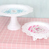 Weiße Spitze-Dessertstand mit blauem Blumen- und rosa Spitzenteller auf karierter rosa Tischdecke.