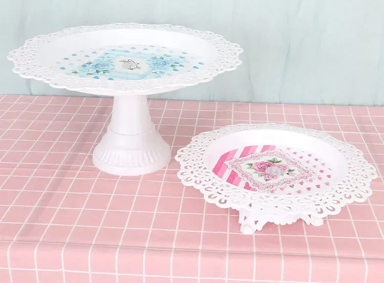 Weiße Spitze-Dessertstand mit blauem Blumen- und rosa Spitzenteller auf karierter rosa Tischdecke.