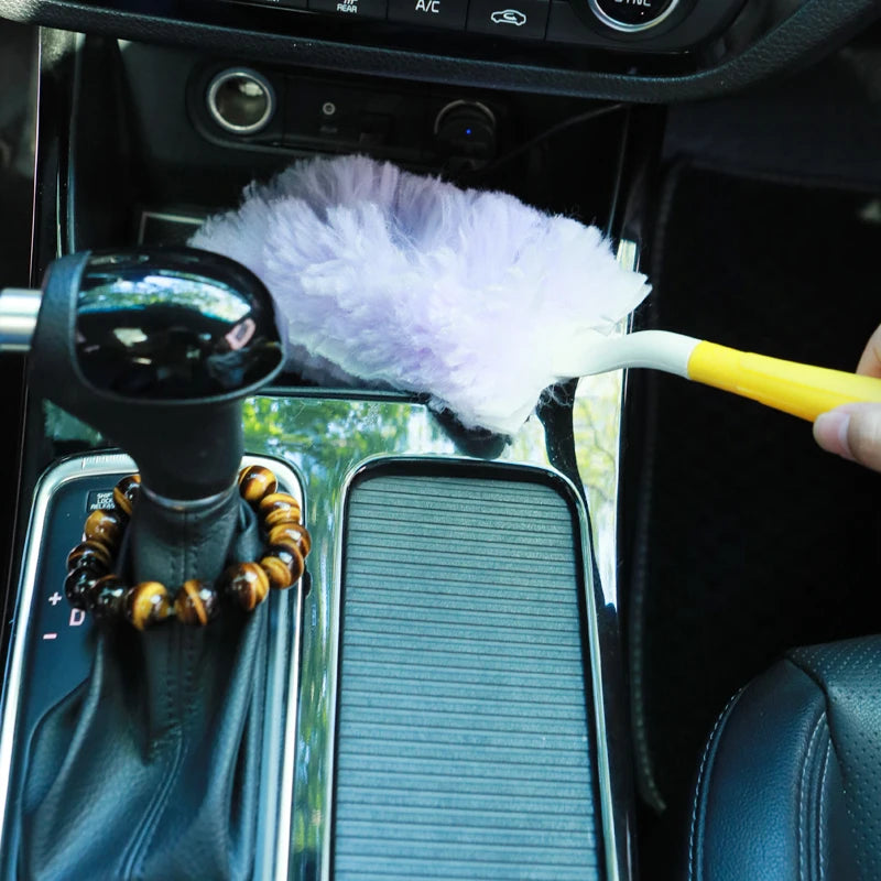A hand is using a purple feather duster to clean the interior of a car, visible through the windshield.