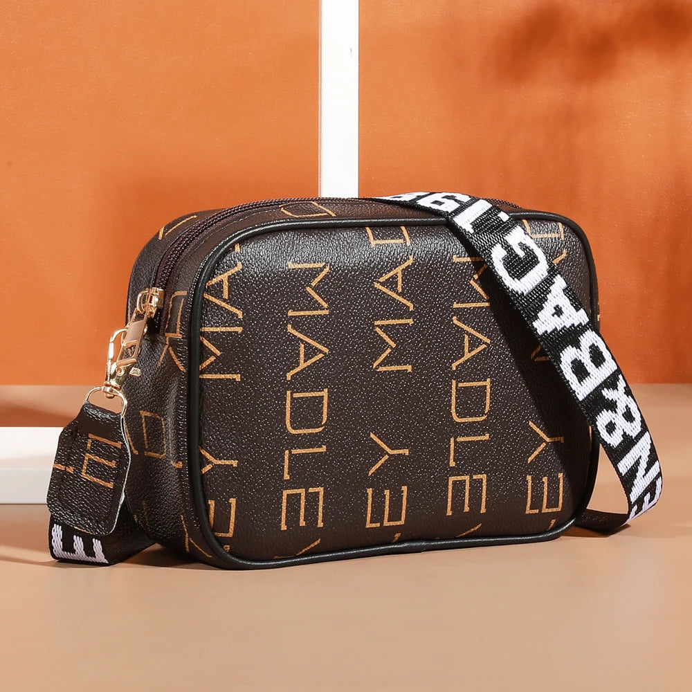 A brown leather compact crossbody bag with gold hardware and black and white striped strap resting on a light surface.