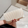 A hand holds a light beige pleated clutch bag with a chain strap against a bright background.