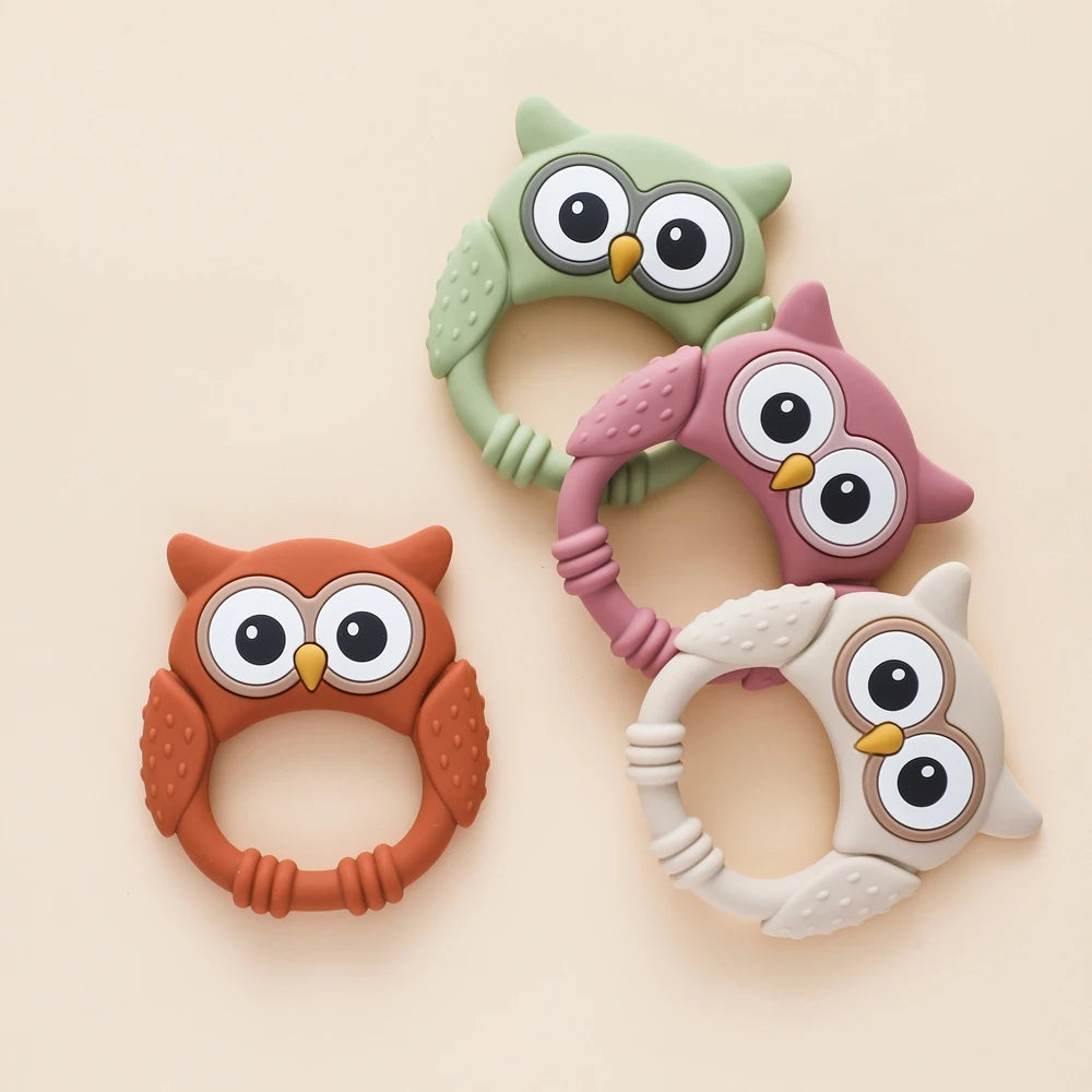 Three silicone owl-shaped teethers and rattles in green, pink, and beige tones.