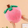 A pink strawberry plush throw pillow with green leaves is being held by a hand.