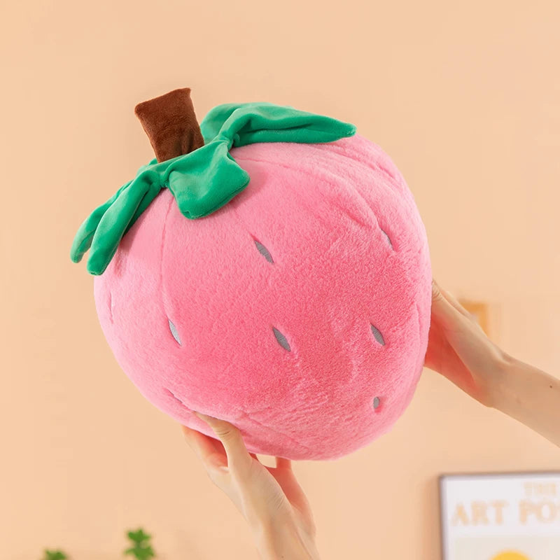 A pink strawberry plush throw pillow with green leaves is being held by a hand.
