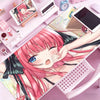 A close-up of a pink-haired anime character illustration on a mouse pad or desk mat, surrounded by stationery and gadgets.