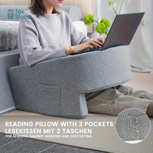 A person is using a gray memory foam reading pillow with two pockets for various activities.