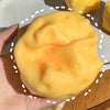 A hand holds a large, smooth, yellow squeeze playful cheese fidget toy against a white background.