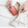 Hands place a compact, reddish-brown leak-proof travel soap box into a light-colored fabric pouch.