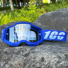 Blue motocross goggles with a blue strap resting on a dark rock outdoors.