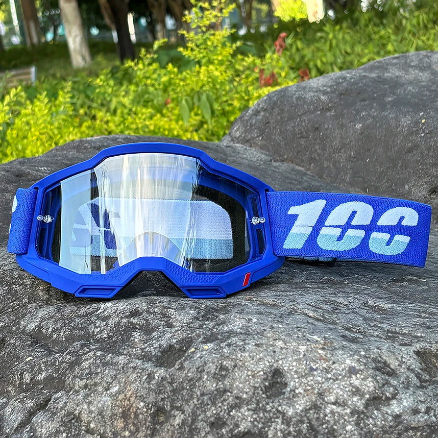 Blue motocross goggles with a blue strap resting on a dark rock outdoors.