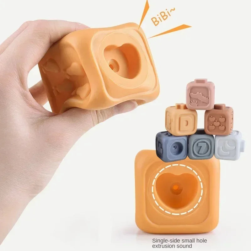 A hand holds an orange silicone building block with a heart-shaped hole, next to several small, colored silicone blocks.