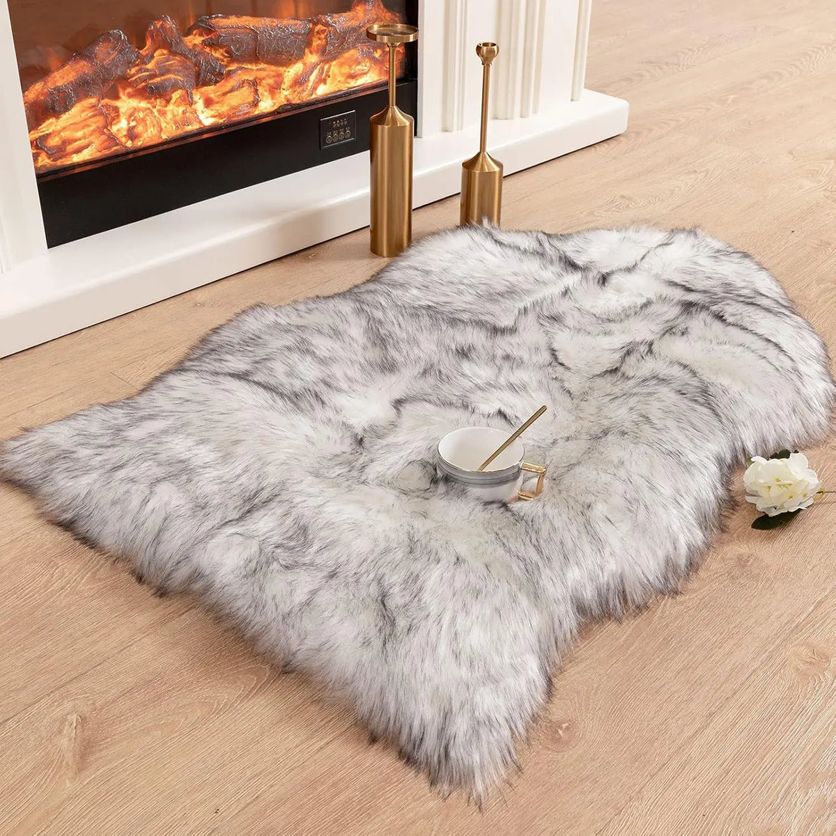 Un tapis en fausse fourrure grise et moelleuse est exposé sur un plancher en bois à côté d'une cheminée et de chandeliers dor