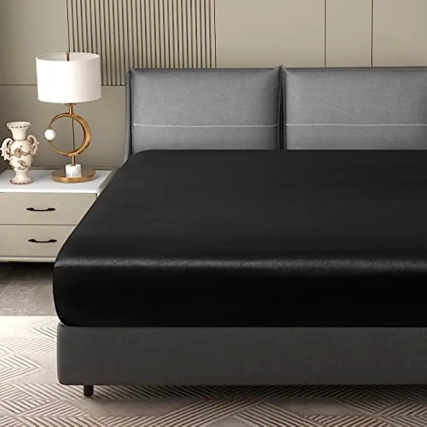 A black luxury satin fitted sheet is displayed on a bed with gray upholstered headboards and bedside table.