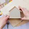 A hand is placing a window frame stencil onto a cardboard house facade for DIY craft projects.