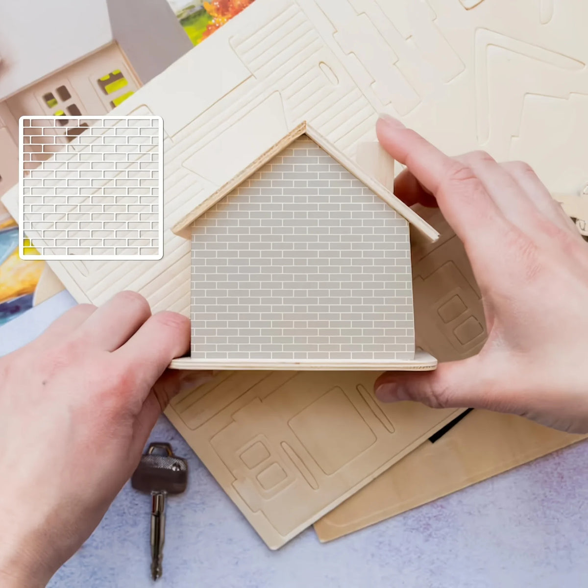 A hand is placing a window frame stencil onto a cardboard house facade for DIY craft projects.