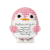 A pink crocheted penguin plush with black eyes and an orange beak, holding a white sign.