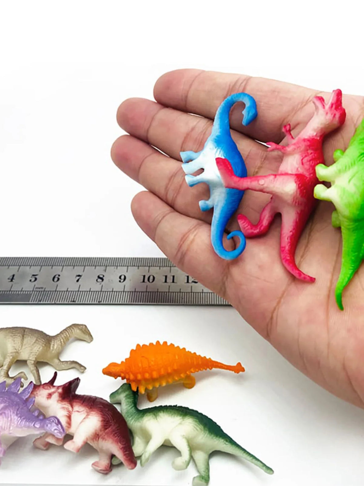 A hand holds a set of ten mini dinosaur figurines, including a blue dragon, pink tyrannosaurus, and various green and purple 