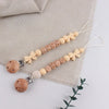 A string of wooden silicone pacifier clips with baby names attached, laid on a beige linen fabric.