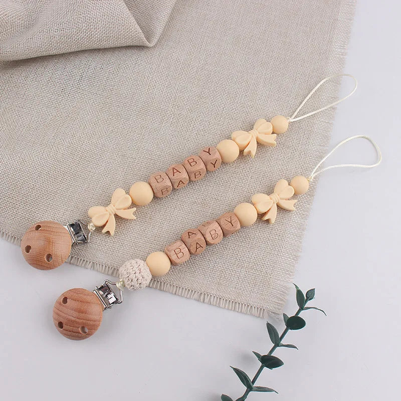 A string of wooden silicone pacifier clips with baby names attached, laid on a beige linen fabric.