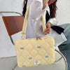 A light yellow quilted laptop shoulder bag with a fabric strap is shown next to a laptop and a metal chair.