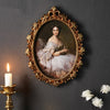 An ornate gold-framed oil painting featuring a woman in white lace dress against a dark gray wall.