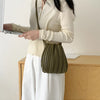A woman holds a olive green pleated faux leather crossbody bag while wearing a cream sweater and white skirt.