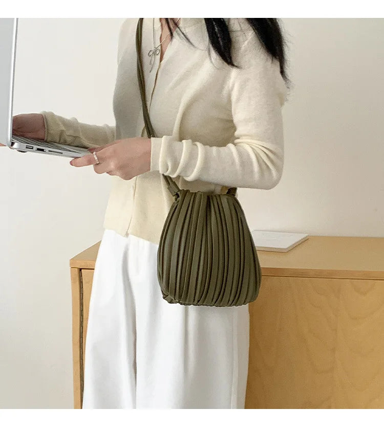 A woman holds a olive green pleated faux leather crossbody bag while wearing a cream sweater and white skirt.