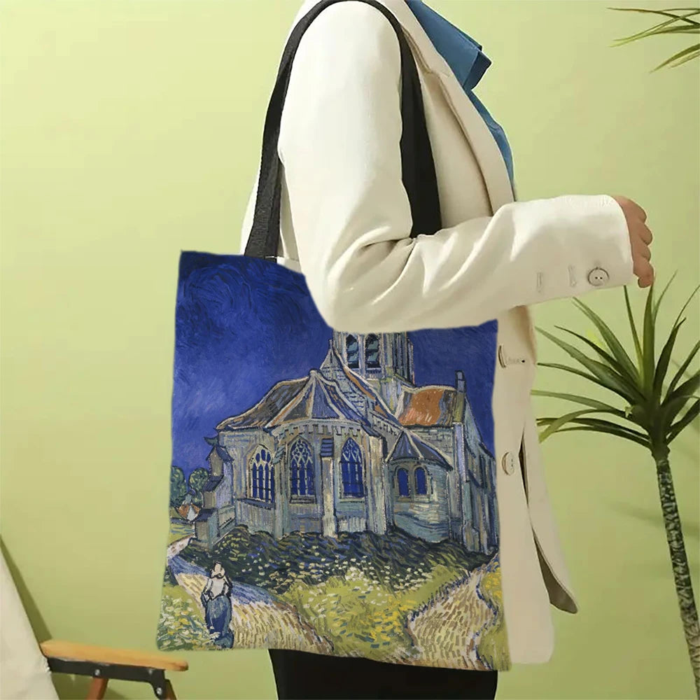 A person holds a canvas tote bag featuring a painting of a church building under a blue sky.