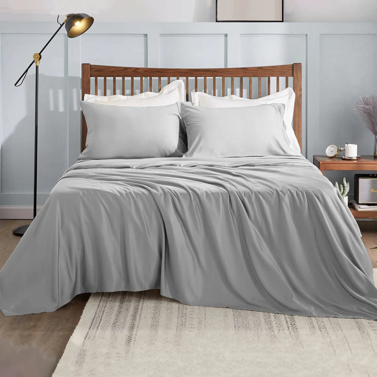 Une chambre avec un lit en bois, des draps blancs et une couette grise sur un tapis clair.