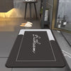 A rectangular dark gray bath mat with white lettering and a light gray border is displayed on a modern bathroom floor.