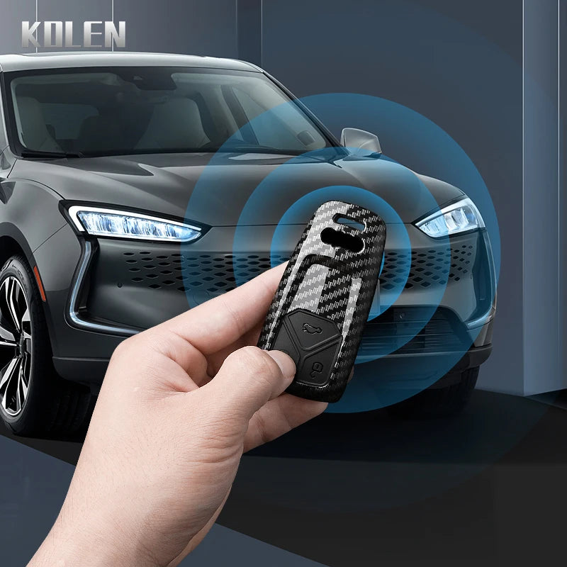 A hand holds a carbon fiber style key fob cover for an Audi car in front of a dark vehicle.