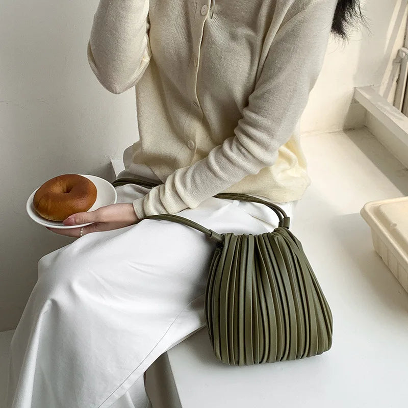 A person wearing a cream-colored top and white pants holds an olive green pleated faux leather crossbody bag.